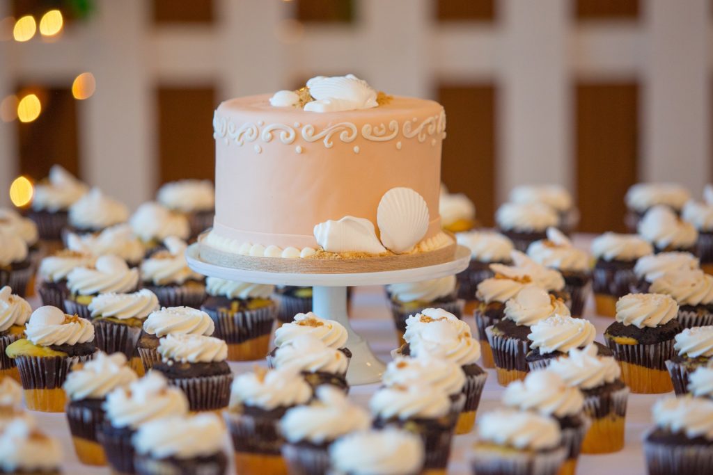 how to charge for cake delivery - A pale pink wedding cake on a pedastool surrounded by cupcakes.